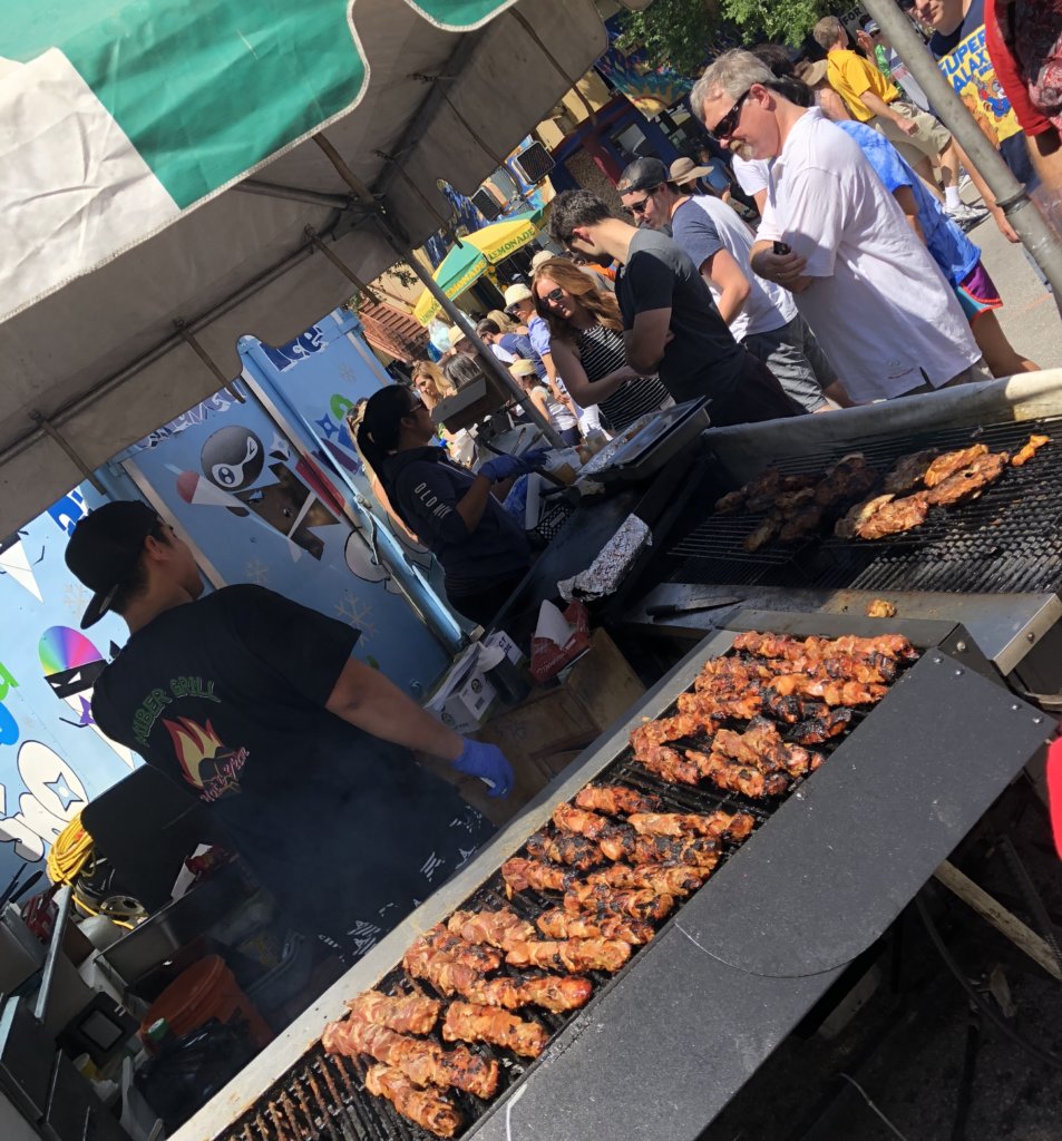Meat kebobs on a barbeque with people on the street celebrating a festival.