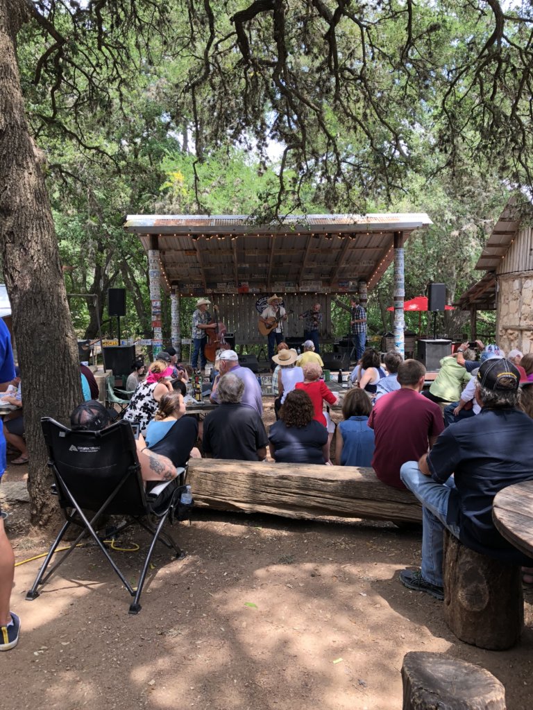 Large, sprawling, Texas oak, country band playing on a stage and people sitting in chairs listening to the music.