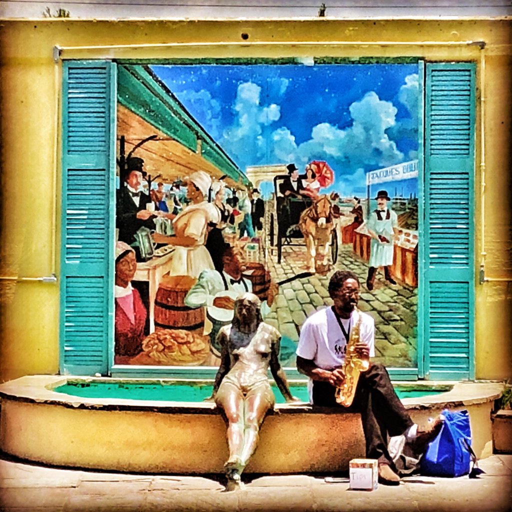 Man playing a trombone sits on a fountain beside a statue. There is a tip jar at his feet. A painting is in the background depicting street life in New Orleans a century ago.