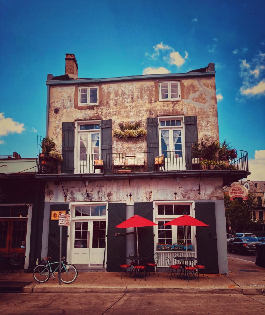 An old two-story building that serves as a house and a pizza kitchen on the main floor. An old, blue bicycle sits out front as well as two red umbrellas that cover tables for guests to sit at. The building is old and dilaputaed but beautiful.