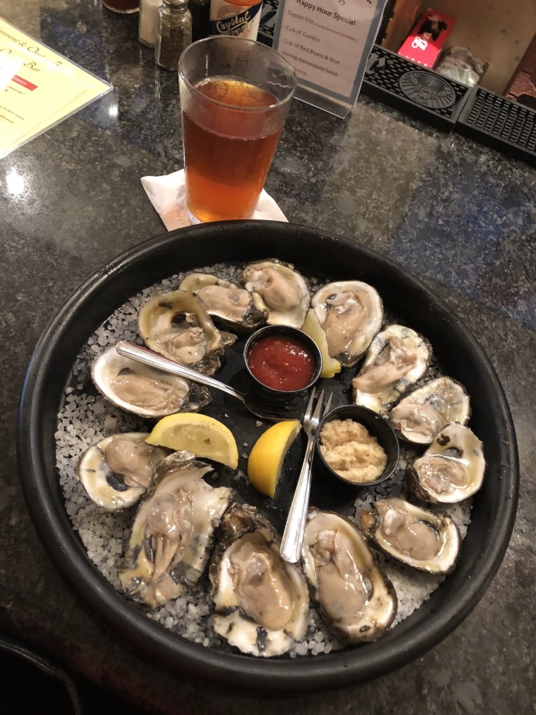 A black plate with a bedding of salt rocks with twelve raw oysters served in the shell. Lemon wedges garnish. Served with seafood sauce and horseradish.