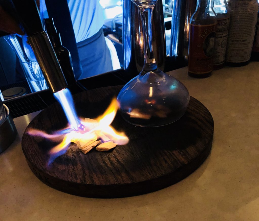 Cedar chips are lit on fire with a small torch, then a martini glass is placed overtop to put out the flames and generate smoke. While the glass smokes, a concoction containing rum, vermouth and bitters is shaken over ice.