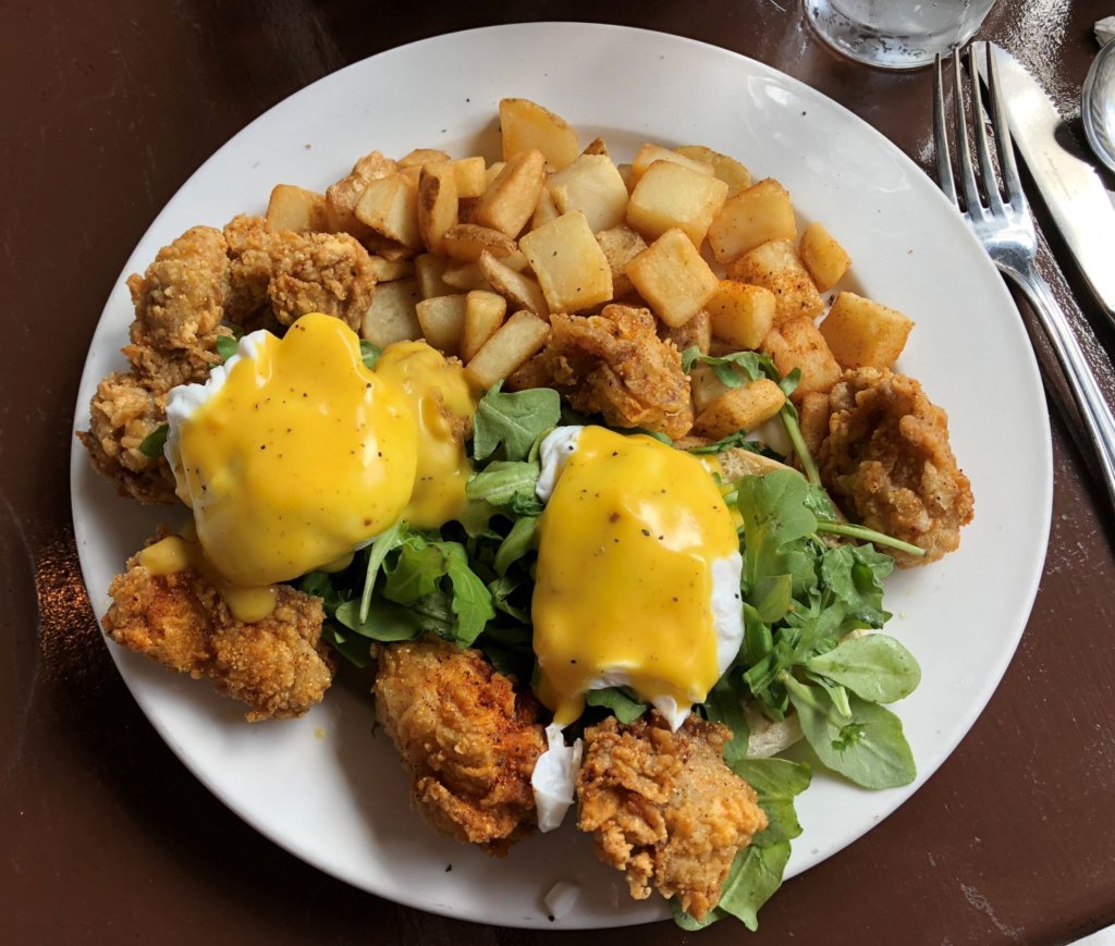 A breakfast dish serving an abundance of battered, fried oysters with poached eggs, hollandaise sauce, arugula and breakfast potatoes.