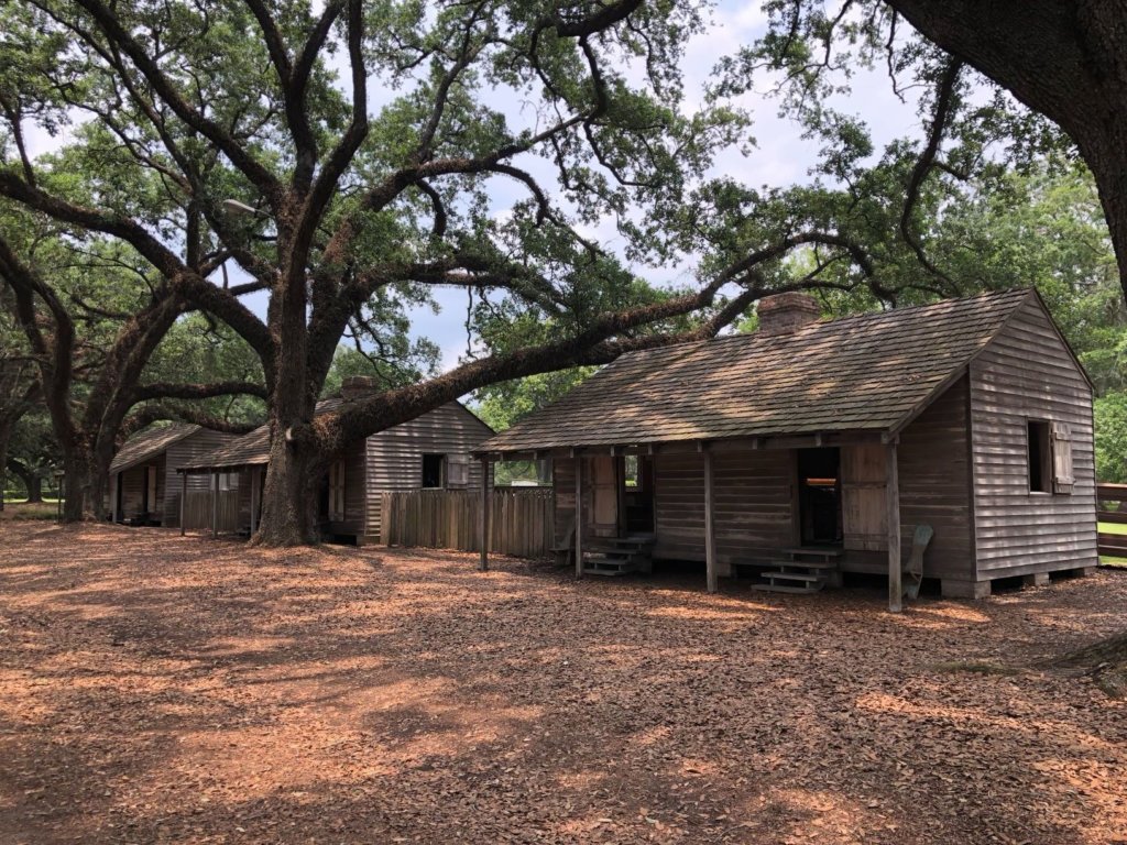 Three plain, wooden shacks underneath the shade of oak trees that were used as slave quarters in the southern United States.