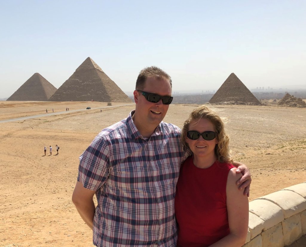 Two happy tourists stand in front of the Pyramids of Giza and pose for a photographer.