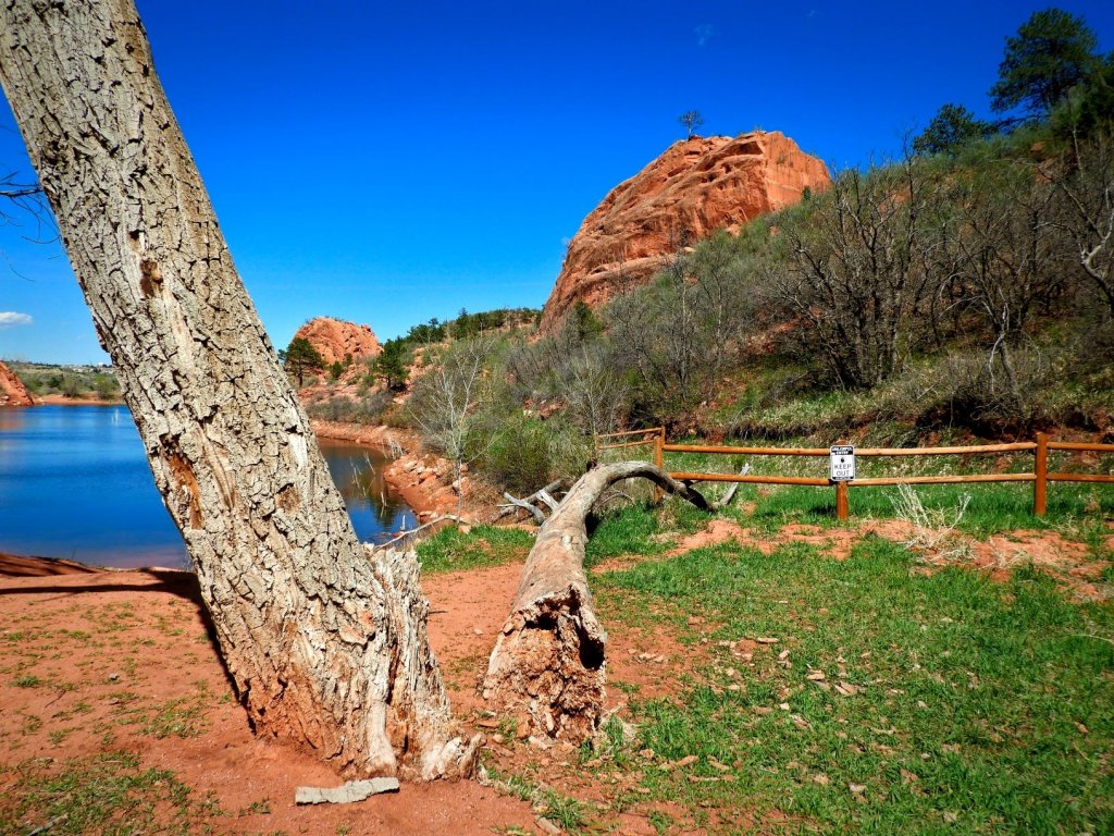 a blue lake, red rocks and dirt with fallen trees and a fence